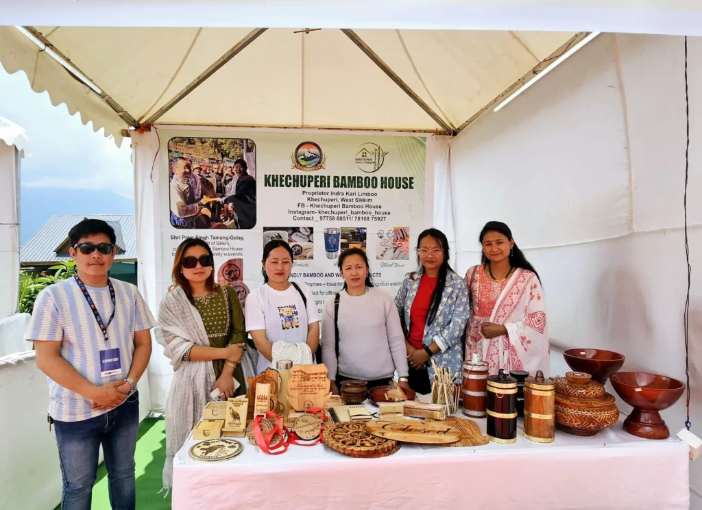 Group of local artisans from Sikkim standing at the Khechuperi Bamboo House exhibition stall, showcasing handcrafted bamboo products including bowls, bottles, trophies, souvenirs, and eco friendly utility items at a cultural fair in West Sikkim.