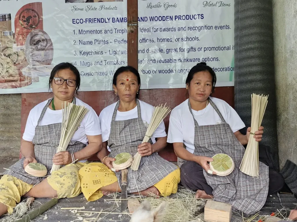 Three women artisans from Sikkim sitting together while handcrafting bamboo products, holding bamboo strips and woven bases, demonstrating traditional eco friendly bamboo weaving techniques at a local handicraft showcase.