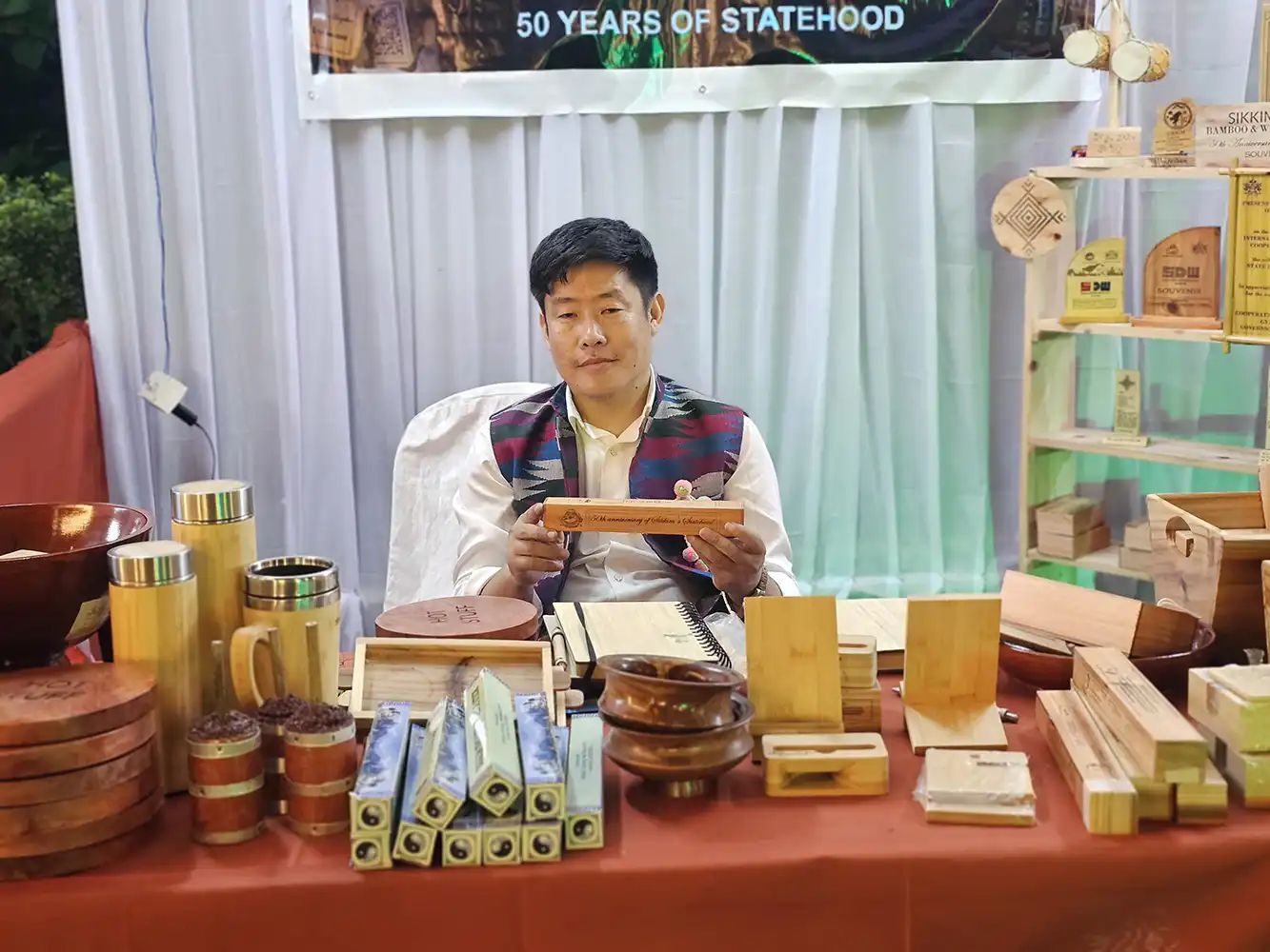 Indra Kari Limboo, founder of Khechuperi Bamboo House, seated at an exhibition stall in Sikkim, holding a handcrafted bamboo product and showcasing eco friendly bamboo and wooden handicrafts including bottles, bowls, mementos, stationery, and souvenirs.