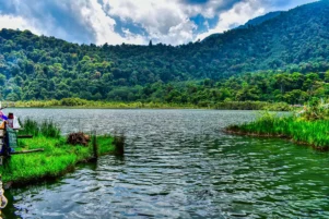 Scenic view of Khechuperi Lake in West Sikkim, surrounded by dense green forested hills and clear water under a bright sky, reflecting the natural beauty and sacred landscape of Sikkim.