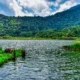 Scenic view of Khechuperi Lake in West Sikkim, surrounded by dense green forested hills and clear water under a bright sky, reflecting the natural beauty and sacred landscape of Sikkim.