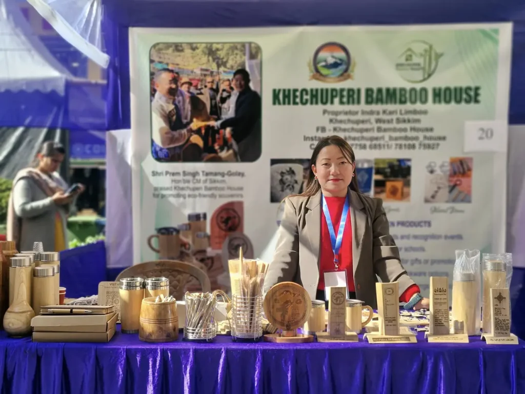 Woman artisan from Sikkim standing at the Khechuperi Bamboo House stall, displaying eco friendly handcrafted bamboo products such as bottles, cups, trophies, stationery, and souvenirs during a local exhibition in West Sikkim.